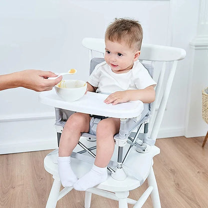 Child sitting in a white high chair with a bowl and spoon, on a light wooden floor.
