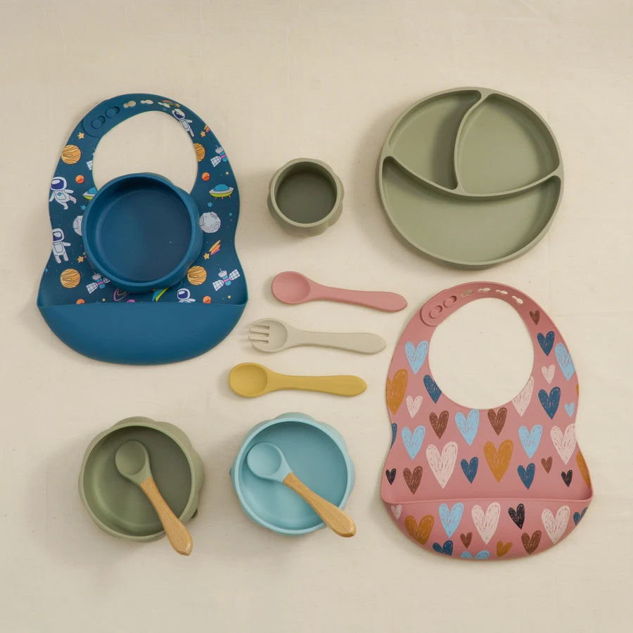 Collection of children's tableware including bibs, plates, bowls, and utensils on a beige background