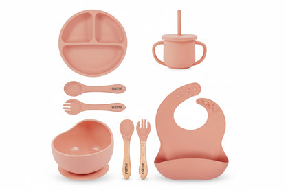 Children's dining set with pink items including a plate, cup, bowl, utensils, and bib on a white background.